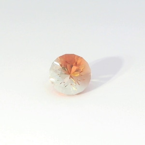 May include: A round, faceted gemstone featuring a color gradient that transitions from a warm, saturated orange at the top to a pale, translucent white at the bottom. The stone has a brilliant cut with multiple triangular facets.