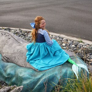 Puede incluir: Una ni&ntilde;a vestida de sirena se sienta en una estatua de ballena. Lleva un vestido azul con una cola verde y un lazo azul en el pelo.
