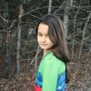 Peut inclure: Une personne portant un costume avec un haut vert &agrave; manches longues, un gilet bleu et dor&eacute; &agrave; motifs, une ceinture rouge et une jupe cr&egrave;me. L'arri&egrave;re-plan pr&eacute;sente une for&ecirc;t avec des arbres d&eacute;nud&eacute;s.