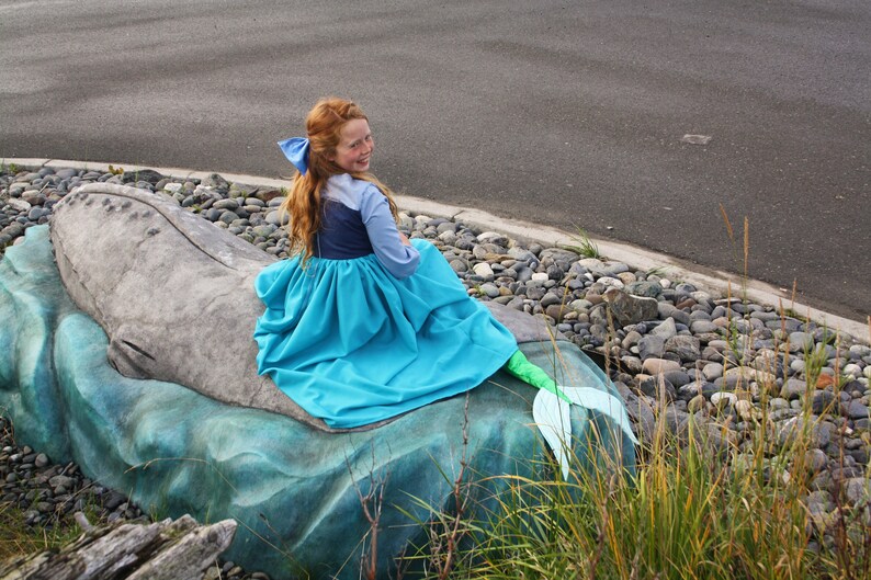 Puede incluir: Una ni&ntilde;a vestida de sirena se sienta en una estatua de ballena. Lleva un vestido azul con una cola verde y un lazo azul en el pelo.