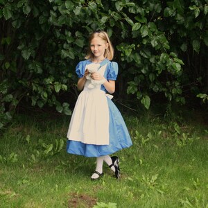 Puede incluir: Una ni&ntilde;a vestida como Alicia en el Pa&iacute;s de las Maravillas, con un vestido azul y blanco con un delantal blanco, sostiene un animal de peluche blanco. Est&aacute; de pie en un &aacute;rea cubierta de hierba con follaje verde en el fondo.