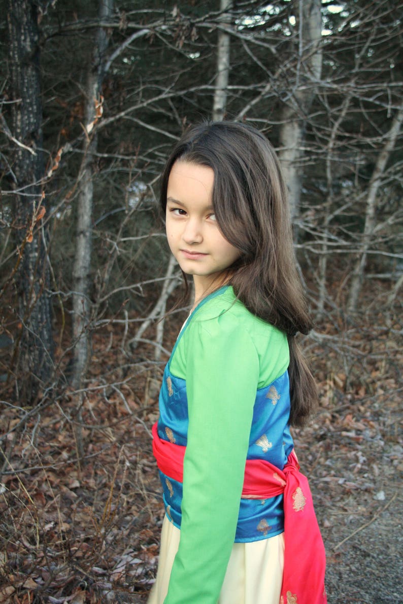 Peut inclure: Une personne portant un costume avec un haut vert &agrave; manches longues, un gilet bleu et dor&eacute; &agrave; motifs, une ceinture rouge et une jupe cr&egrave;me. L'arri&egrave;re-plan pr&eacute;sente une for&ecirc;t avec des arbres d&eacute;nud&eacute;s.