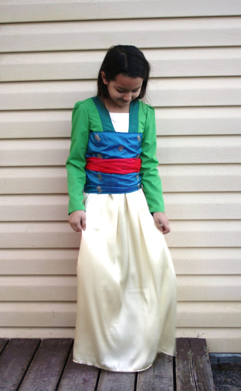 Peut inclure: Une robe de costume verte, bleue et rouge. La robe pr&eacute;sente un haut vert &agrave; manches longues, un corsage bleu avec des accents dor&eacute;s, une ceinture rouge et une longue jupe cr&egrave;me. Le costume est probablement pour un enfant.