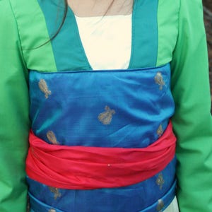 Peut inclure: Un costume vert et bleu avec des accents dor&eacute;s. Le costume comprend une veste verte avec une ceinture bleue et rouge. Le tissu bleu pr&eacute;sente des motifs floraux dor&eacute;s. Le costume est probablement pour une pi&egrave;ce de th&eacute;&acirc;tre ou un cosplay.