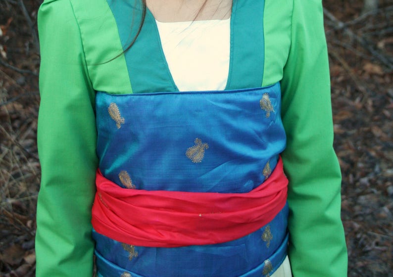 Peut inclure: Un costume vert et bleu avec des accents dor&eacute;s. Le costume comprend une veste verte avec une ceinture bleue et rouge. Le tissu bleu pr&eacute;sente des motifs floraux dor&eacute;s. Le costume est probablement pour une pi&egrave;ce de th&eacute;&acirc;tre ou un cosplay.