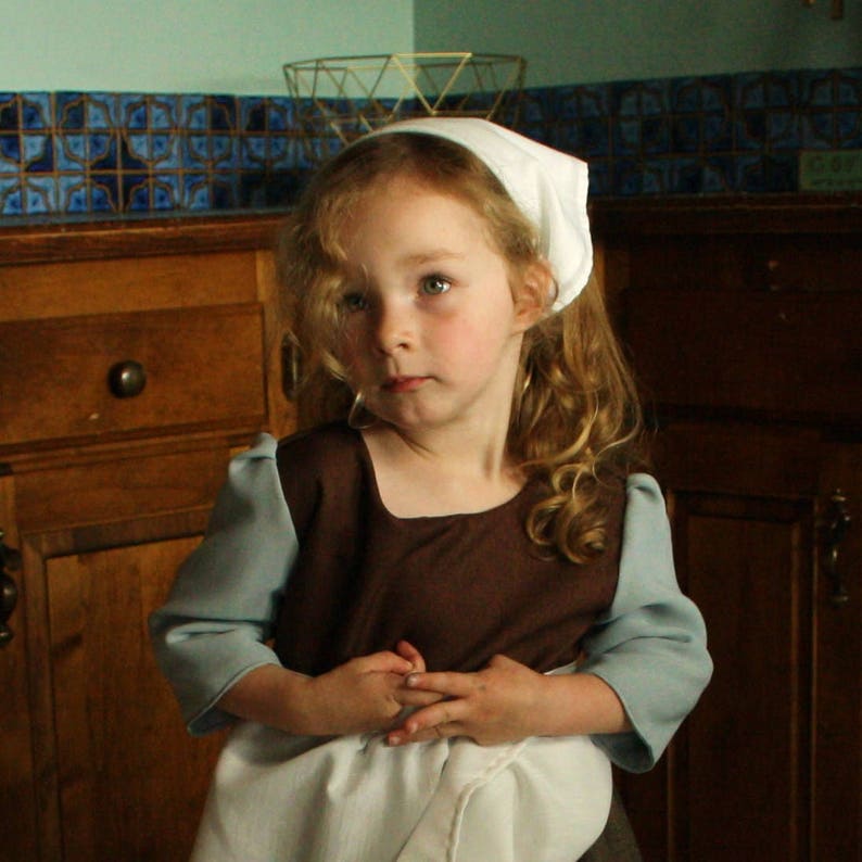May include: A young girl wearing a brown dress with a white apron and a white headband. She has long, curly blonde hair and is looking to the side. The girl is standing in front of a wooden cabinet.