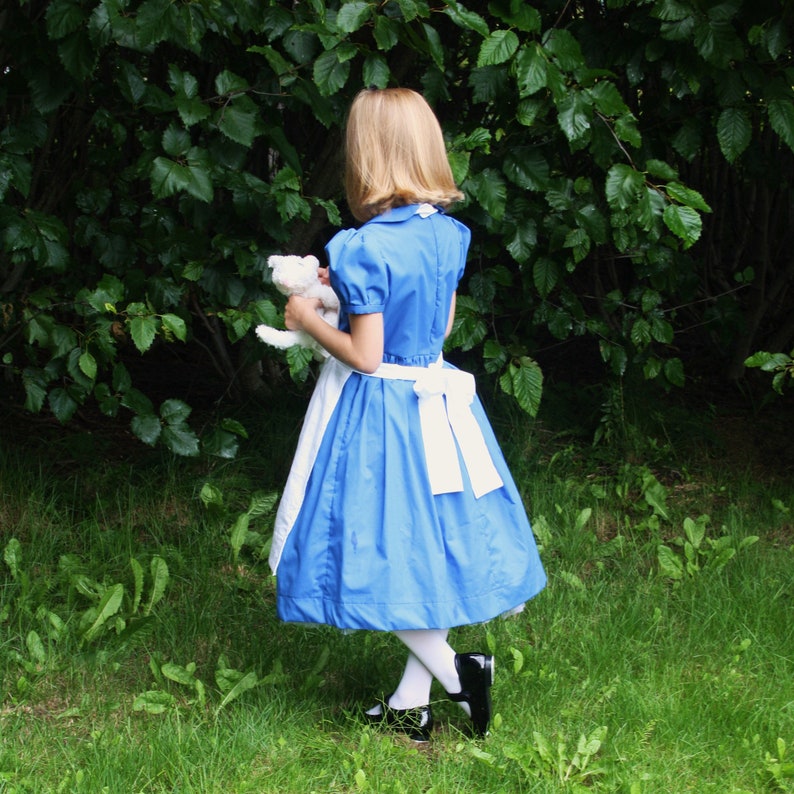 Puede incluir: Una ni&ntilde;a vestida como Alicia en el Pa&iacute;s de las Maravillas, con un vestido azul y blanco con un delantal blanco y una faja blanca. Lleva un conejo de peluche blanco en sus brazos y est&aacute; de pie frente a un arbusto verde.