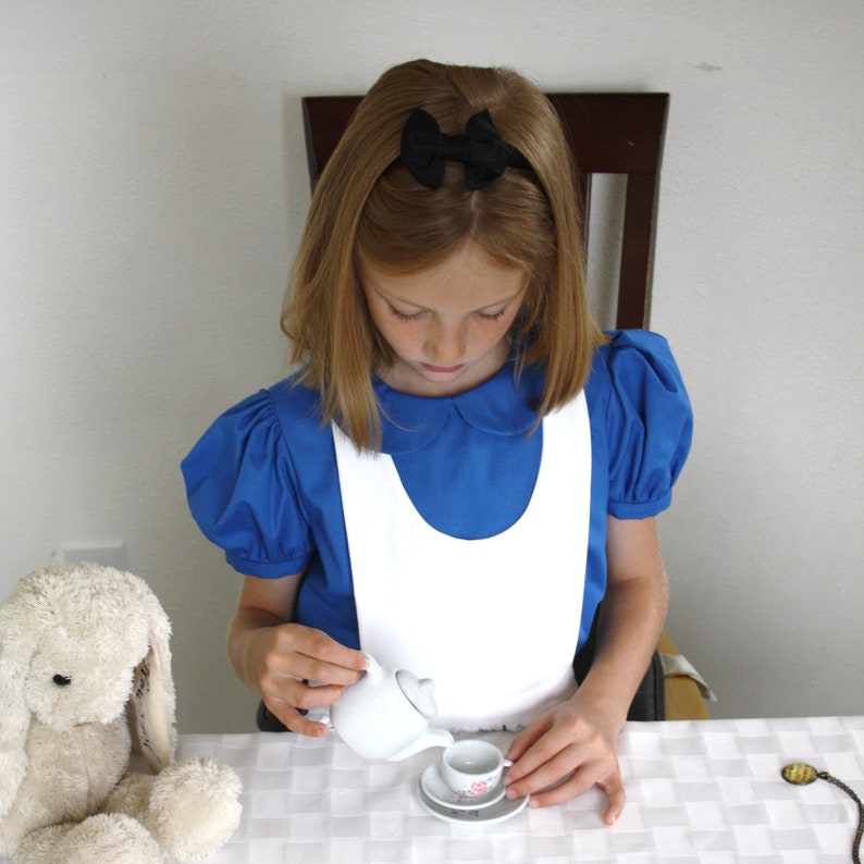 Puede incluir: Una ni&ntilde;a vestida con un vestido azul y blanco vierte t&eacute; de una tetera blanca en una peque&ntilde;a taza de t&eacute; blanca. La taza de t&eacute; est&aacute; sobre un platillo blanco con un dise&ntilde;o floral rosa. La ni&ntilde;a lleva un lazo negro en el pelo.