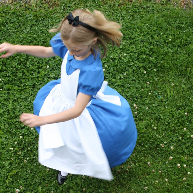 Puede incluir: Una ni&ntilde;a peque&ntilde;a con un vestido azul y blanco con un delantal blanco y un lazo negro en el pelo. Est&aacute; dando vueltas en un campo de hierba.