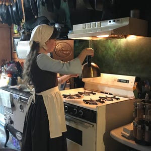 May include: A woman wearing a white bonnet and a black dress with a white apron is standing in a kitchen. She is holding a copper pot lid over a white gas stove with four burners. The stove has a black oven below it. There are many cast iron skillets hanging from the ceiling above the stove.