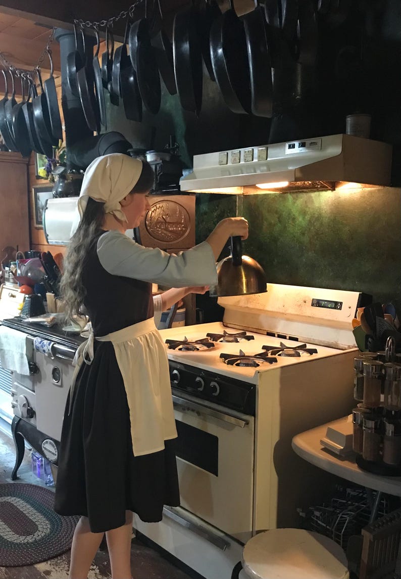 May include: A woman wearing a white bonnet and a black dress with a white apron is standing in a kitchen. She is holding a copper pot lid over a white gas stove with four burners. The stove has a black oven below it. There are many cast iron skillets hanging from the ceiling above the stove.