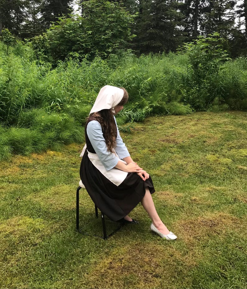 May include: A woman wearing a white bonnet and a black and white dress sits on a black stool in a grassy field.