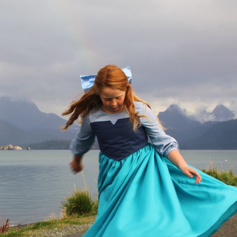 Puede incluir: Una ni&ntilde;a peque&ntilde;a con un disfraz de princesa azul y turquesa con un lazo azul en el pelo. Est&aacute; de pie frente a una masa de agua con monta&ntilde;as en el fondo.