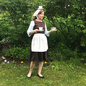 May include: A woman dressed as a maid is holding two teacups and saucers. She is wearing a brown dress with a white apron and a white head scarf. She is standing in a grassy area with green trees in the background.