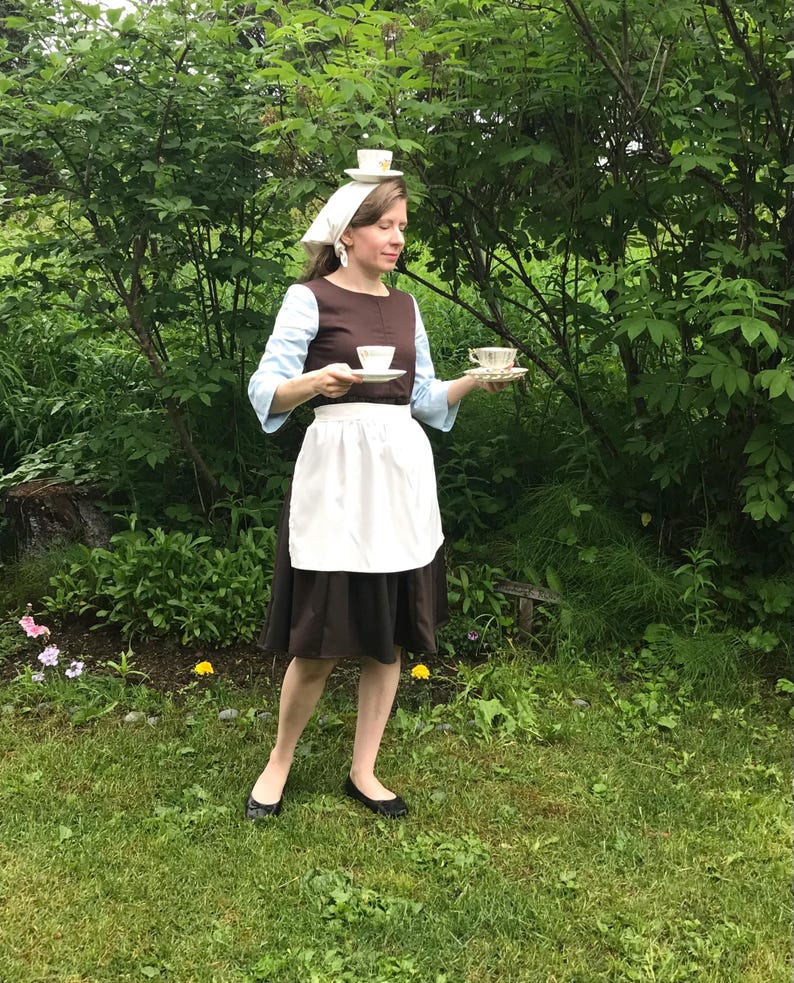 May include: A woman dressed as a maid is holding two teacups and saucers. She is wearing a brown dress with a white apron and a white head scarf. She is standing in a grassy area with green trees in the background.