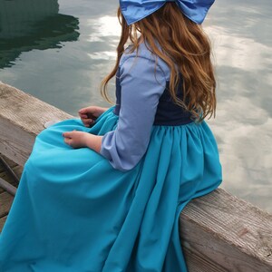 Puede incluir: Una ni&ntilde;a peque&ntilde;a con un vestido azul y azul marino con un gran lazo azul en el pelo. Est&aacute; sentada en un muelle de madera con una masa de agua detr&aacute;s de ella.