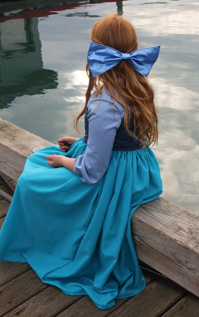 Puede incluir: Una ni&ntilde;a peque&ntilde;a con un vestido azul y azul marino con un gran lazo azul en el pelo. Est&aacute; sentada en un muelle de madera con una masa de agua detr&aacute;s de ella.