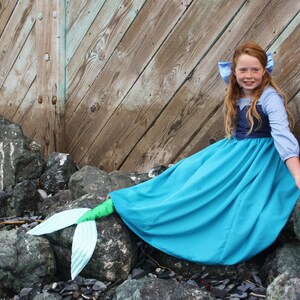 Puede incluir: Una ni&ntilde;a vestida de sirena se sienta en las rocas junto a una pared de madera. Lleva un vestido azul y morado con una cola azul.