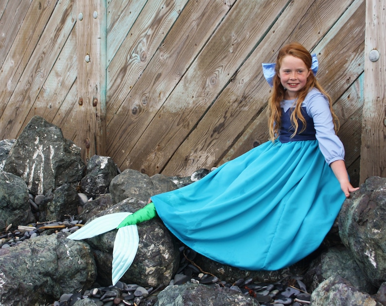 Puede incluir: Una ni&ntilde;a vestida de sirena se sienta en las rocas junto a una pared de madera. Lleva un vestido azul y morado con una cola azul.