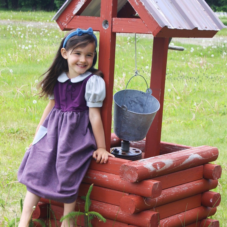 以下が含まれることがあります： 青いヘッドバンドをした紫と白のドレスを着た少女が、ロープにバケツがぶら下がった木の井戸のそばに座っています。