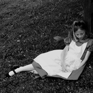 Puede incluir: Una ni&ntilde;a vestida con un vestido blanco con una cinta roja y un delantal blanco se sienta en la hierba. Lleva una diadema y tiene el pelo recogido hacia atr&aacute;s. Est&aacute; mirando algo en su regazo.