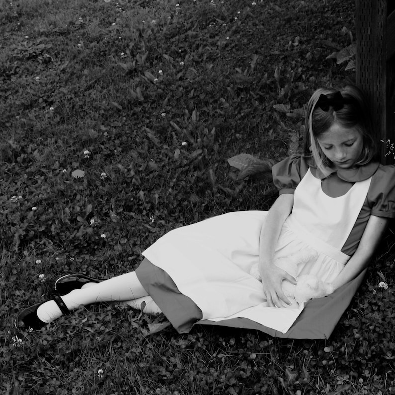 Puede incluir: Una ni&ntilde;a vestida con un vestido blanco con una cinta roja y un delantal blanco se sienta en la hierba. Lleva una diadema y tiene el pelo recogido hacia atr&aacute;s. Est&aacute; mirando algo en su regazo.