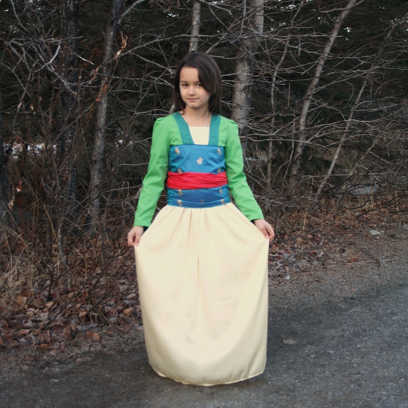 Peut inclure: Une robe de costume avec un haut vert, un corsage turquoise avec des d&eacute;tails dor&eacute;s, une ceinture rouge et une longue jupe cr&egrave;me. Les manches sont longues et vertes. La robe est port&eacute;e par une personne devant des arbres.