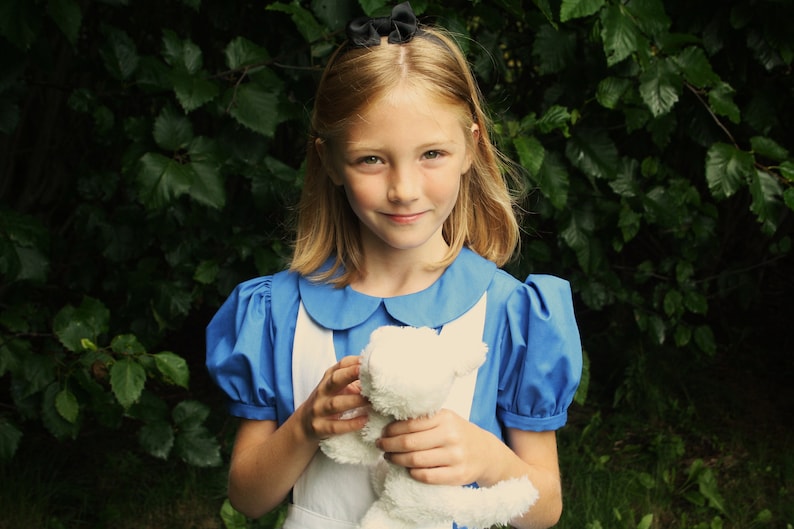 Puede incluir: Una ni&ntilde;a peque&ntilde;a con un vestido azul con cuello blanco y un lazo blanco en el pelo. Est&aacute; sosteniendo un animal de peluche blanco en sus manos. La ni&ntilde;a est&aacute; de pie frente a un fondo verde frondoso.