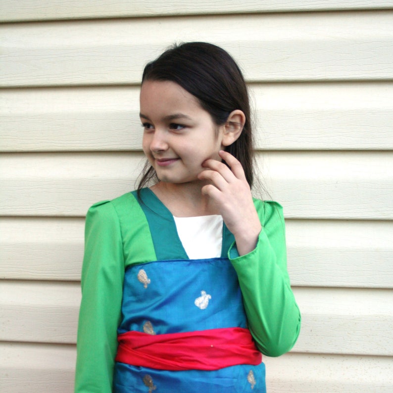 Peut inclure: Robe de costume verte et bleue avec des manches longues et une ceinture rouge. La robe a des accents dor&eacute;s et une encolure blanche. Le costume est port&eacute; par une personne aux cheveux fonc&eacute;s.