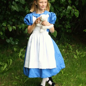 Puede incluir: Una ni&ntilde;a vestida como Alicia en el Pa&iacute;s de las Maravillas, con un vestido azul y blanco con un delantal blanco, sostiene un animal de peluche blanco. Est&aacute; de pie en una zona cubierta de hierba con vegetaci&oacute;n verde en el fondo.