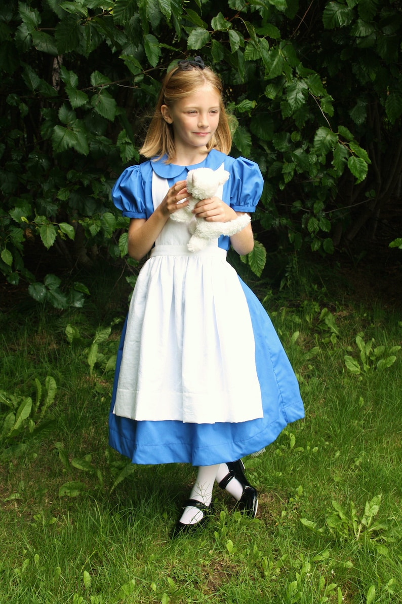 Puede incluir: Una ni&ntilde;a vestida como Alicia en el Pa&iacute;s de las Maravillas, con un vestido azul y blanco con un delantal blanco, sostiene un animal de peluche blanco. Est&aacute; de pie en una zona cubierta de hierba con vegetaci&oacute;n verde en el fondo.