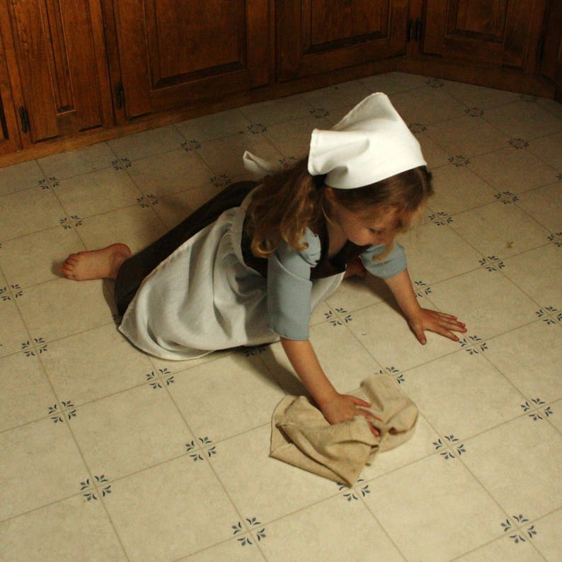 May include: A young person wearing a white cap and a white and brown dress is kneeling on a tiled floor and cleaning with a cloth.