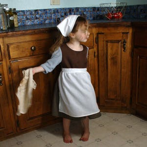May include: A young person wearing a brown and white dress with a white bonnet and a blue shirt is holding a white cloth in a kitchen. The kitchen has wooden cabinets and a tiled backsplash.