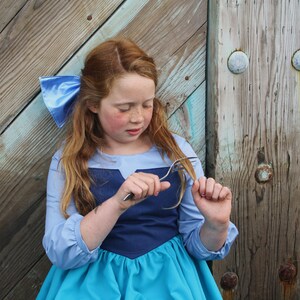 Puede incluir: Una ni&ntilde;a peque&ntilde;a con un vestido azul y turquesa con un lazo azul en el pelo. Est&aacute; sosteniendo un tenedor en su mano y lo est&aacute; mirando.