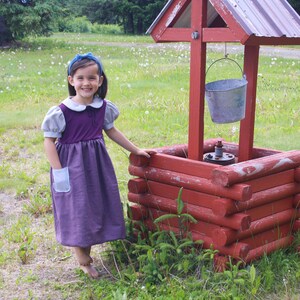 以下が含まれることがあります： 青いヘッドバンドをした紫と灰色のドレスを着た若い女の子が、赤い木製の井戸の横に立っています。井戸にはバケツがぶら下がっています。