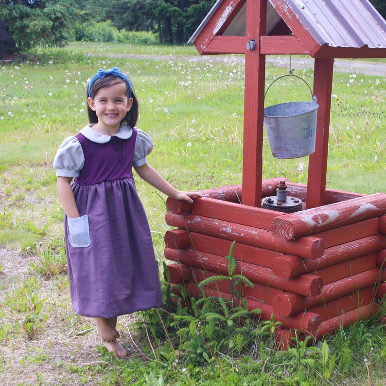 以下が含まれることがあります： 青いヘッドバンドをした紫と灰色のドレスを着た若い女の子が、赤い木製の井戸の横に立っています。井戸にはバケツがぶら下がっています。