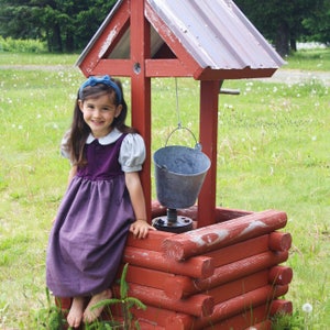 以下が含まれることがあります： 白い襟と青いリボンをつけた紫色のドレスを着た若い女の子が、ロープにバケツがぶら下がった木製の井戸の前に立っています。