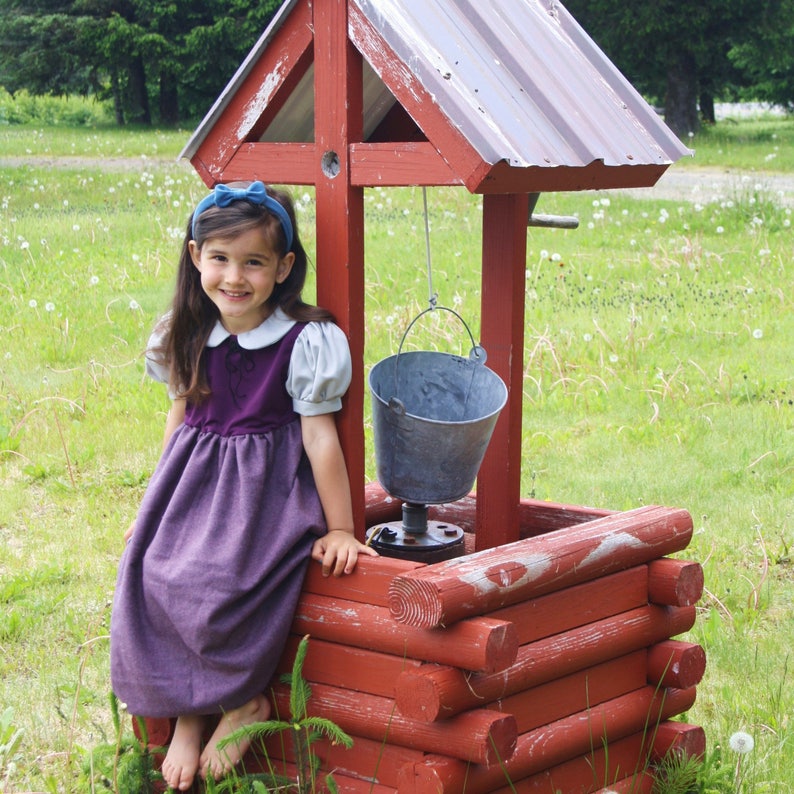 以下が含まれることがあります： 白い襟と青いリボンをつけた紫色のドレスを着た若い女の子が、ロープにバケツがぶら下がった木製の井戸の前に立っています。