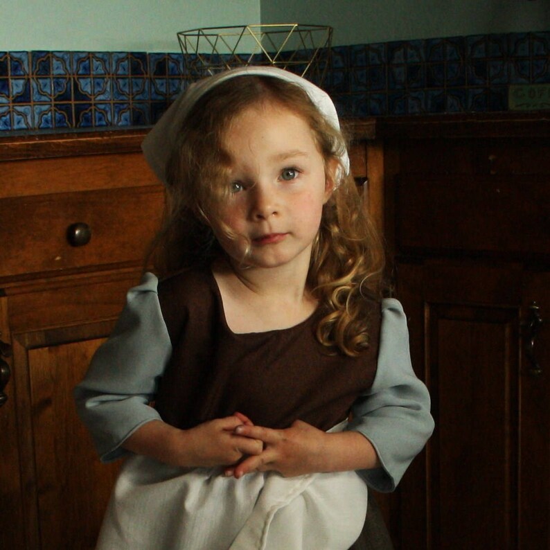 May include: A young girl with long blonde hair wears a white bonnet and a brown dress with blue sleeves. She is standing in front of a wooden cabinet.
