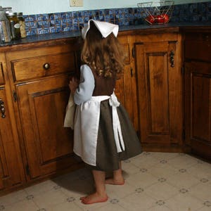 May include: A young girl wearing a white and blue dress with a white apron and a white headband. She is standing in a kitchen with wooden cabinets and a tiled backsplash.