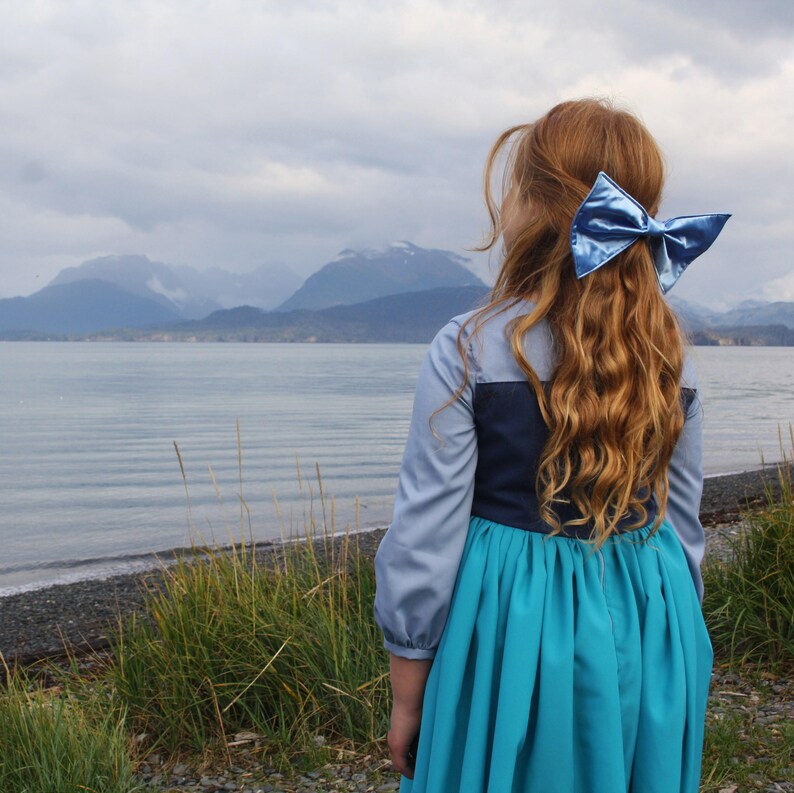 Puede incluir: Una ni&ntilde;a con el pelo largo y rizado rojo que lleva un vestido azul y turquesa con un lazo de sat&eacute;n azul en el pelo. Est&aacute; de pie en una playa con vistas a las monta&ntilde;as y el agua en el fondo.