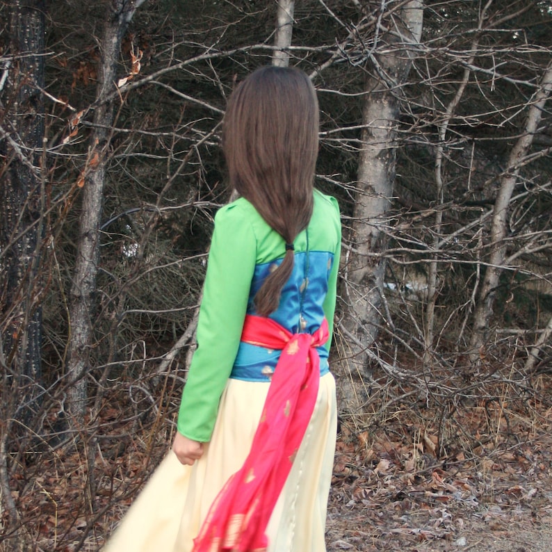 Peut inclure: Haut vert &agrave; manches longues avec un corsage &agrave; motifs bleus et dor&eacute;s et une longue jupe cr&egrave;me. Une &eacute;charpe rouge est nou&eacute;e &agrave; la taille. Les longs cheveux bruns de la personne sont tress&eacute;s.