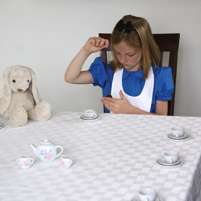 Puede incluir: Una ni&ntilde;a peque&ntilde;a con un vestido azul y un delantal blanco se sienta en una mesa con un mantel blanco. Est&aacute; sosteniendo un objeto peque&ntilde;o y redondo en su mano. Hay varias tazas de t&eacute; y platillos en la mesa, as&iacute; como una tetera y un conejito de peluche.