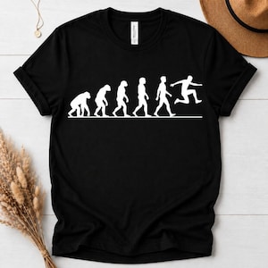 May include: A black short-sleeved t-shirt featuring a white graphic print of the human evolution sequence. The design shows the progression from a primate to a human, ending with a figure in a dynamic jumping pose, all aligned on a horizontal line.