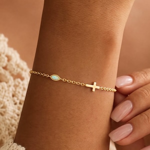May include: A delicate gold chain bracelet featuring a small, horizontal cross charm and a single, oval-shaped iridescent opal stone set in a gold bezel. The bracelet is constructed with fine, uniform links and secures with a simple clasp mechanism.