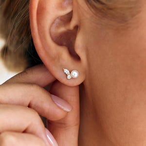 May include: A gold stud earring featuring a single white pearl paired with two small, clear, marquise-cut stones arranged in a cluster. The earring is positioned on the earlobe, showcasing a delicate, asymmetrical design with a polished metal finish.