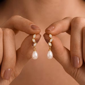 May include: A pair of gold-toned earrings featuring a vertical row of three marquise-cut clear stones leading to a single white teardrop-shaped pearl pendant. The earrings are constructed with a delicate gold metal setting that holds the stones and pearl.
