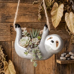 May include: A gray ceramic sloth-shaped hanging planter suspended by jute twine. The planter features a white face with brown eye markings and a black nose. It is filled with a variety of green and light-colored succulent plants, including a spiky haworthia.