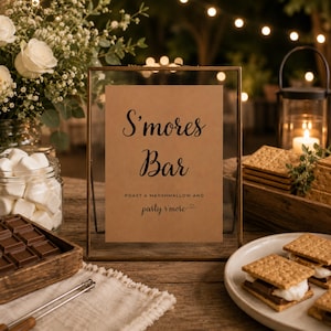 May include: A rectangular glass frame with a brass border displaying a tan sign that reads S'mores Bar, Roast a marshmallow and party s'more. The sign is surrounded by a glass jar of white marshmallows, a wooden tray of chocolate, and graham crackers.