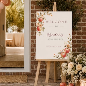 Op de afbeelding: Een rechthoekig wit welkomstbord op een houten ezel met aquarelillustraties van rode appels en herfstbladeren. De tekst luidt: WELCOME TO Kendra's BABY SHOWER OCTOBER 25TH, 2026. Het ontwerp bevat florale accenten in de hoeken van het bord.
